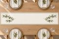 Ivory olive branch table runner with a fine green double border on a wooden dining table, set with white plates and folded napkins.