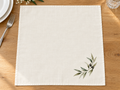 Ivory olive branch napkin laid open on a wooden table, showing a watercolor olive sprig with dark olives in the lower-right corner.